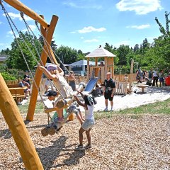Auf einer Mehrgenerationschaukel sitzen mehrere Kinder und ein Kind schubst an. Dahinter ist eine Banksitzgruppe und eine Rutsche und ein Holzturm zu sehen. Es ist ein großer Sandkasten um die Rutsche und Holzspäne um die Schaukel. Auf dem Spielplatz sind viele Menschen und im Hintergrund viele Bäume.
