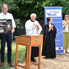 Hinter einem Rednerpult steht eine Frau in schwarzen Leggins und weißem Shirt und ein Mann mit dunkler Hose und weißem Hemd und Brille und halten einen großen schwarzen Scheck mit weißer Schrift in den Händen. Daneben steht ein Mann im weißem Diakongewand und daneben eine Frau, die ihr schwarzes Pfarrgewand trägt. Dahinter ist ein Roll-up-Banner und zwei Frauen stehen im Sommerkleid daneben.