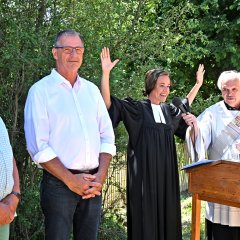 Vor einem Rednerpult steht ein Mann im weißem Diakongewand und hält ein Mikrofon vor eine Frau, die ihr schwarzes Pfarrgewand trägt und die Hände nach oben streckt, um zu segnen. Daneben steht ein Mann mit dunkler Hose und weißem Hemd und Brille und daneben ein Mann mit grüner Hose, weiß-grün karriertem Hemd und ebenso mit Brille.