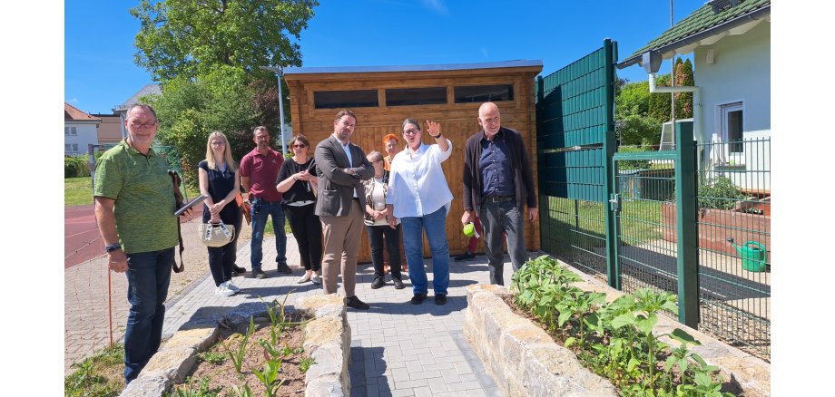 Schulleiterin Katja Dülfer erläutert Landrat Sandro Zehner die Umbaumaßnahmen auf dem Schulgelände der Freiherr-vom-Stein-Schule in Eltville. Hinter zwei langen Hochbeten mit Steinumrandung und Bepfalnzung, stehen vier Männer und fünf Frauen vor einer Holzhütte. In der näheren Umgebung sind weitere Häuser erkennbar und grüne Bäume sowie ein blauer Himmel ohne Wolken.