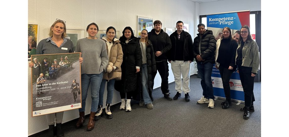 Sieben Frauen und drei Männer stehen vor einem Banner des Kompetenzzentrum Pflege. Die Frau ganz links hält einen Rahmen mit einem Werbeplakat für die Ausstellung "Das Alter in der Karikatur" in der Hand. 