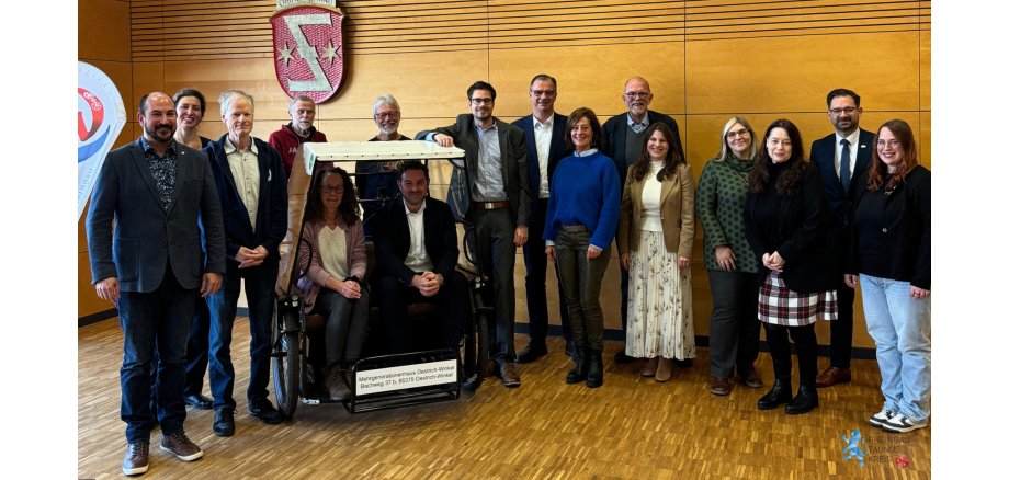 Übergabe OeWi - 1 Vor einer mit Holz verkleideten Wand stehen 8 Männer und 6 Frauen im Halbkreis. Im Halbkreis ist eine Fahrradrikscha für zwei Personen mit beigem Dach, in der eine Frau und ein Mann sitzen, integriert. An der Wand hängt ein rotes Wappen mit einem spiegelverkehrtem "Z" und jeweils einem Stern links und rechts davon. Der Boden ist ein Parkettboden mit kleinen Rechtecken.