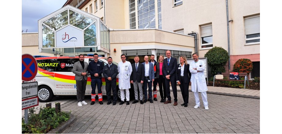 Vor einem beigem Gebäude steht ein Notarztwagen der Malteser und davor stehenneun Männer und zwei Frauen, z.T. im Anzug oder auch Arztkitteln oder Rettungswagenuniform.