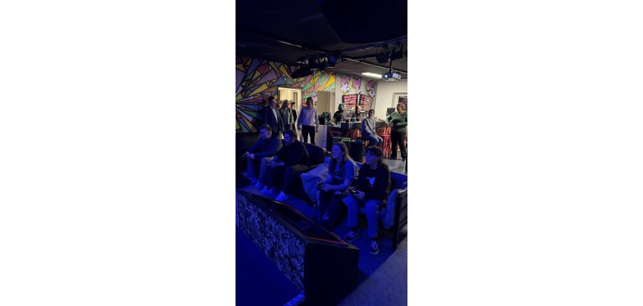 In einem Raum sind Grafitti, lauter bunte Dreiecke mit schwarzen Rahmen und Buchstaben an der Wand. Im Hintergrund ist eine Bar und im Vordergrund zwei Sofas, die mit vier Personen besetzt sind und Gamecontroler in den Händen halten. Es schimmert bläuliches Licht und es sind mehrere Personen im Raum.