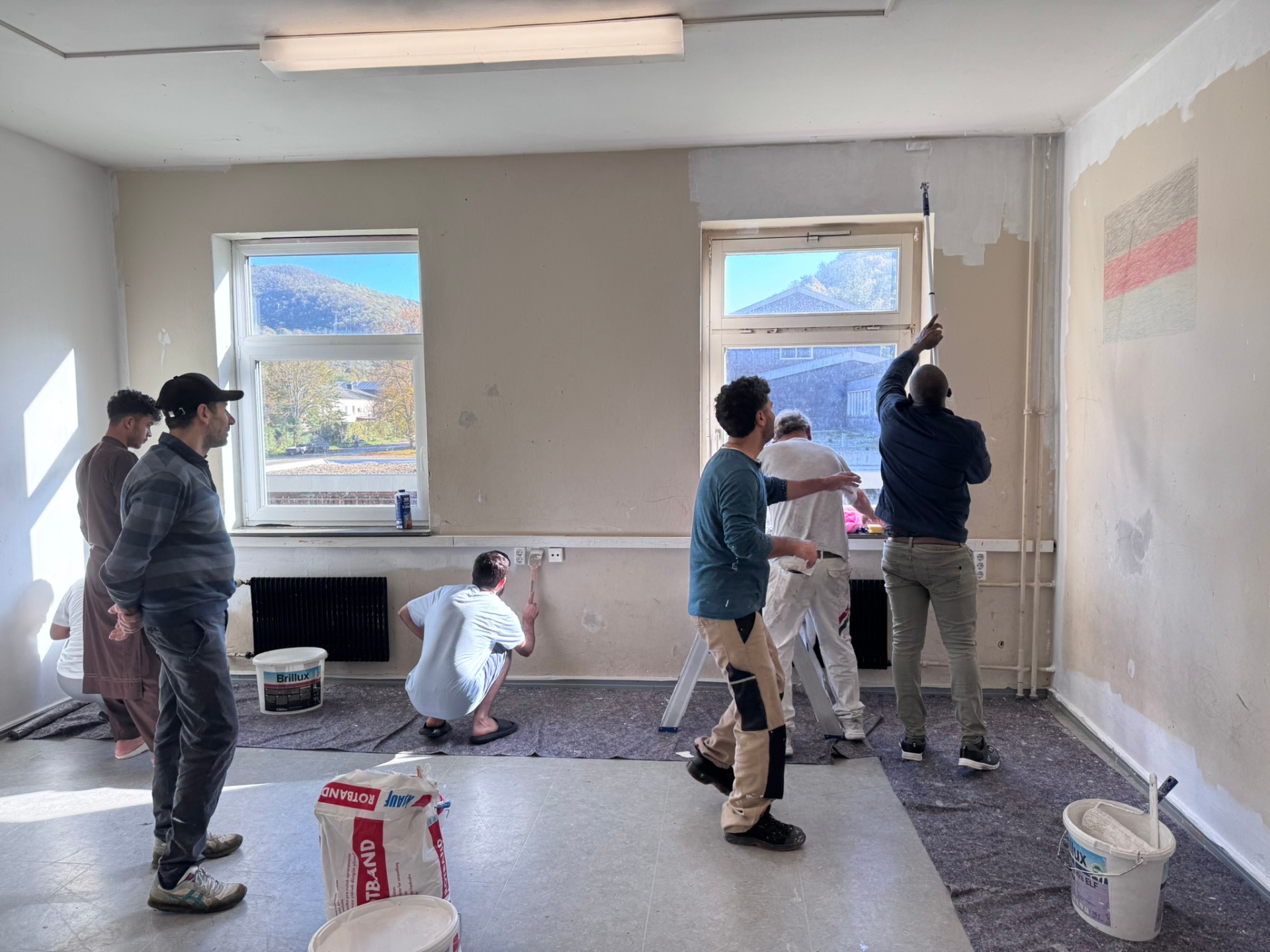 In einem Raum mit zwei Fenstern brennt eine rechteckige Lampe an der Decke und der Boden ist am Rand mit Malerflies ausgelegt. An einer Wand ist eine Deutschlandfahne aufgemalt. Man sieht sieben Männer, die zusammen den Raum mit weißer Farbe streichen.