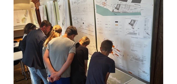 Vor mehreren Pinwänden, auf denene diverse beschriftete Blätter gepinnt wurden, stehen mehrere Kinder und Erwachsene, in Summe 7, die sich vorbeugen um sich etwas anzuschauen.