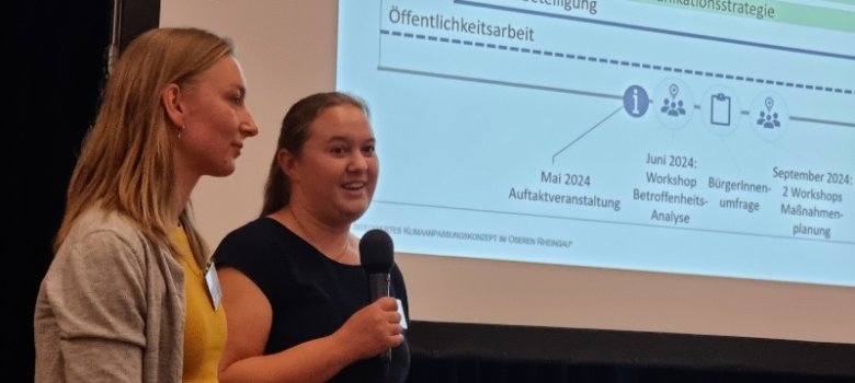 Vor einer großen Leinwand, auf der eine Präsentation läuft, stehen zwei Frauen. Links die Frau hat lange, blonde Haare und ein gelbes Shirt und eine beige Strickjacke an und rechts daneben steht eine Frau mit Mikrophon in der Hand, einem schwarzem T-Shirt und braunen zu einem Pferdeschwanz zusammengebundenen Haaren.