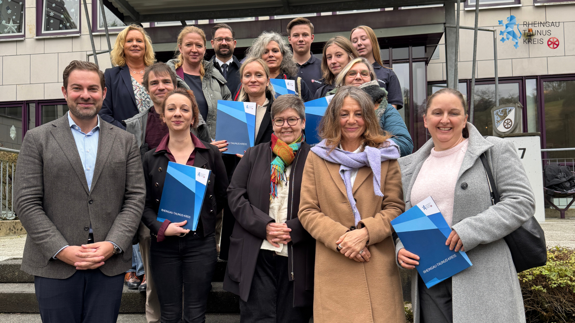 Vor einem Bürogebäude stehen auf Treppenstufen vier Männer und elf Frauen auf unterschidlichen Treppenstufen. Alle tragen WInterjacken. Einige halten blaue Dokumentenmappen in der Hand.