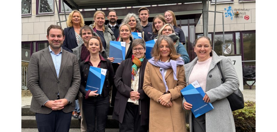 Vor einem Bürogebäude stehen auf Treppenstufen vier Männer und elf Frauen auf unterschidlichen Treppenstufen. Alle tragen WInterjacken. Einige halten blaue Dokumentenmappen in der Hand.