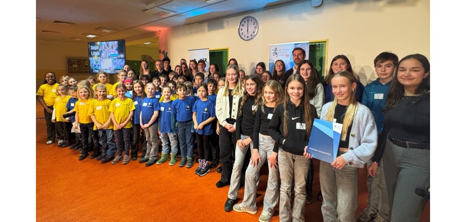 Schulwettbewerb "Miteinander macht Schule" Schülerinnen und Schüler im Hintergrund Leinwand und eine große Bahnhofsuhr an der Wand. Viele Schülerinnen und Schüler tragen gelbe oder blaue T-Shirts.