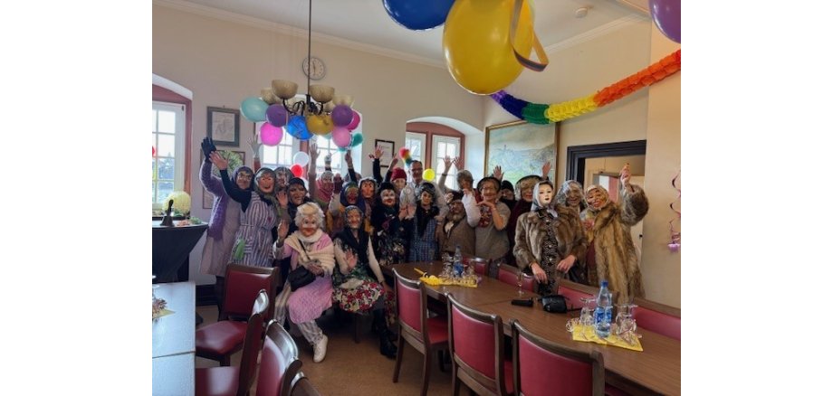 Im dem mit Luftbarlons und Girlanden geschmückten Rathaussaal, steht der Bürgermeister, umringt von 23 Frauen, die als alte Damen verkleidet sind. Sie haben Masken auf und Mäntel oder Kittelschürzen an.