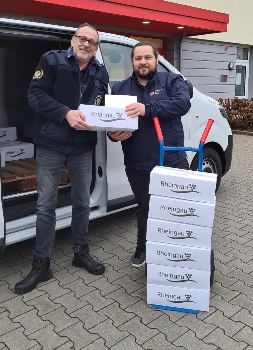 Vor einem weißen Lieferwagen mit geöffneter Schiebetür stehen zwei Männer mit Winterjacken und überreichen sich eine Kiste Wein. Im Lieferwagen liegt eine Palette mit weiteren weißen Weinkisten. Vor dem Lieferwagen steht eine Sackkarre mit sechs weißen Kisten Wein.