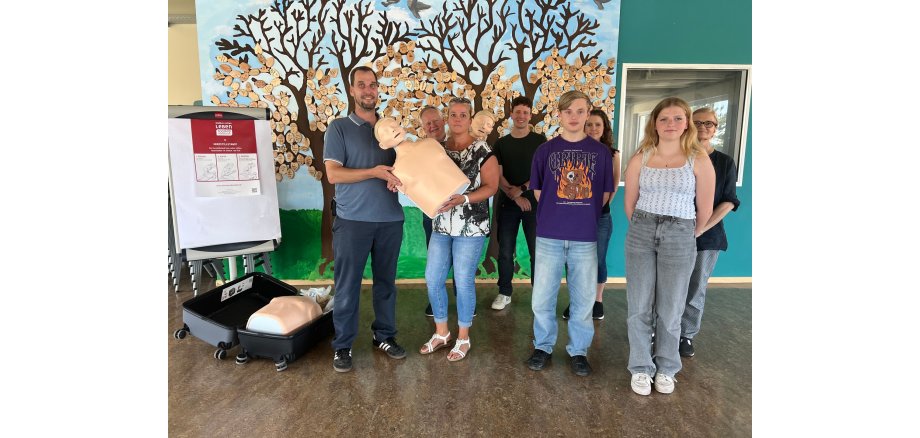 Vor zwei gemalten Bäumen, an denen statt grünen Blätter, hölzerne, beschriftete Blätter hängen, stehen 8 Personen und halten einen Kopf mit Oberkörper einer Trainingspuppe. Vor einem Flipchart mit Plakat, liegt ein offener Koffer mit einer weiteren Trainingspuppe.