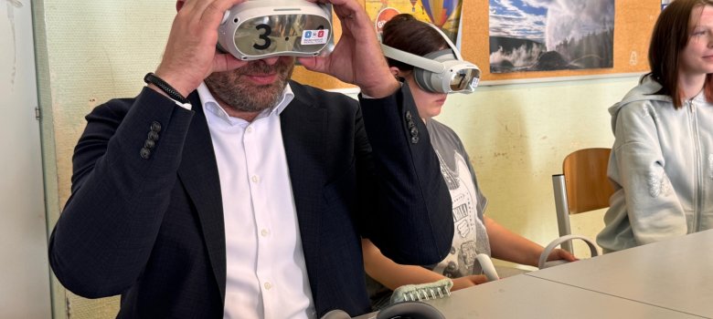 Zwei Schülerinnen und Landrat Zehner sitzen an einem Tisch. An der Wand ist eine Pinnwand mit Plakaten. Ein Mädchen und Landrat Zehner haben eine Headset mit Brille auf und auf dem Tisch liegt eine weitere Brille und Cursor.