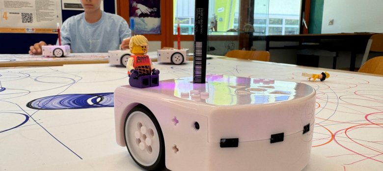 An einem Tisch sitzt ein Junge mit hellblauem Shirt und dunklen Haaren. Hinter ihm ist eine Stellwand mit Plakaten. Der Tisch ist mit weißem Papier ausgelegt. Auf dem Tisch stehen 4 weiße Fahrzeuge mit Rädern, auf denene je ein Stift befestigt ist. Mit dem Stift malt das Fahrzeug Kreise, Linien und Spiralen auf das Papier.