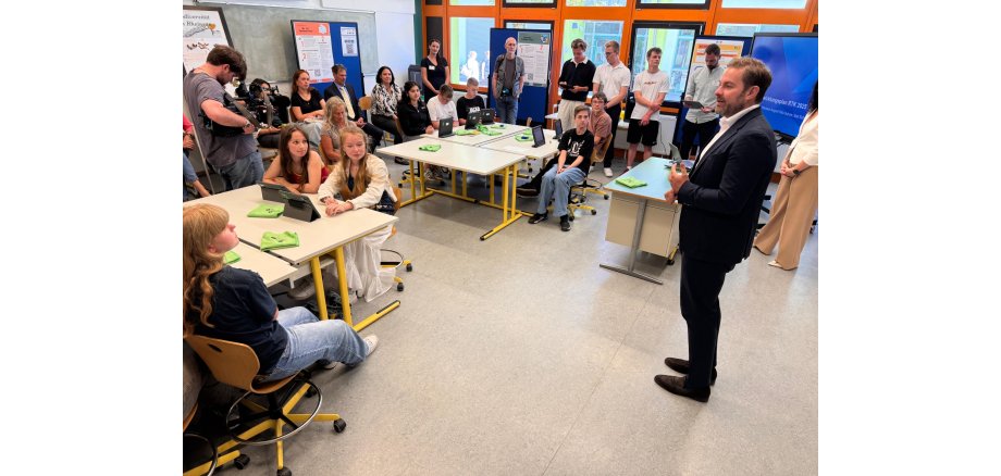 In einem Klassenraum sitzen mehrere Schülerinnen und Schüler an Tischen. Vor ihnen steht jeweils ein Tablett. Außen rum sitzen und stehen weitere Personen. Vor den Wänden und der Fenstefront sind Stellwände mit Plakaten und eine Bildschirm. Vorn steht Landrat Zehner im Anzug und weißem Hemd.