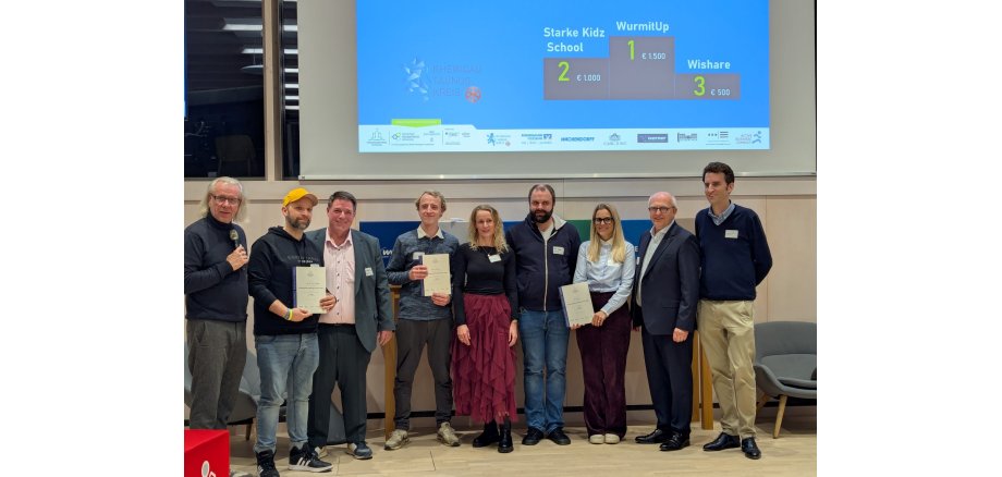 Vor einer großen Leinwand, auf der eine Folie mit drei Säulen, die zeigen, wer welchen Platz belegt hat, abgebildet ist, stehen sieben Männer und zwei Frauen. Zwei Männer und eine Frau halten beige-blaue Urkunden in der Hand.