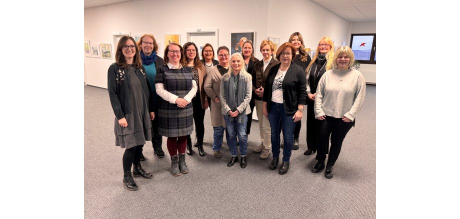 In einem L-förmigem Flur mit Bildern an der Wand und zwei Türen an der einen Seite und einem Fenster mit einer Heizung darunter stehen nebeneinander 13 Frauen.