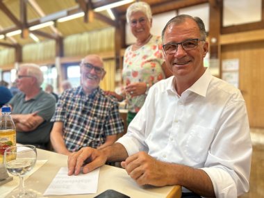 Ein Mann im weißem Hemd, blauer Brille und dunklen Haaren sitzt an einem Tisch. Neben ihm sitzt ein Mann mit Brille und weiß-blau-karriertem Hemd. Dahinter steht eine Damen in weißer Bluse mit binten Blumen und Brille und weißen kurzen Haaren.