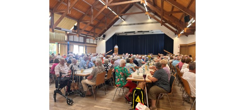 In einem großem Saal, in dem sich eine Bühne mit einem blauem, dicken Bühnenvorhang befindet, steht ein Mann im weißem Hemd am braunem Rednerpult auf der Bühne. Der Saal ist mit Tischen und Stühlen bestückt. Auf den Stühlen sitzen viele Männer und Frauen.