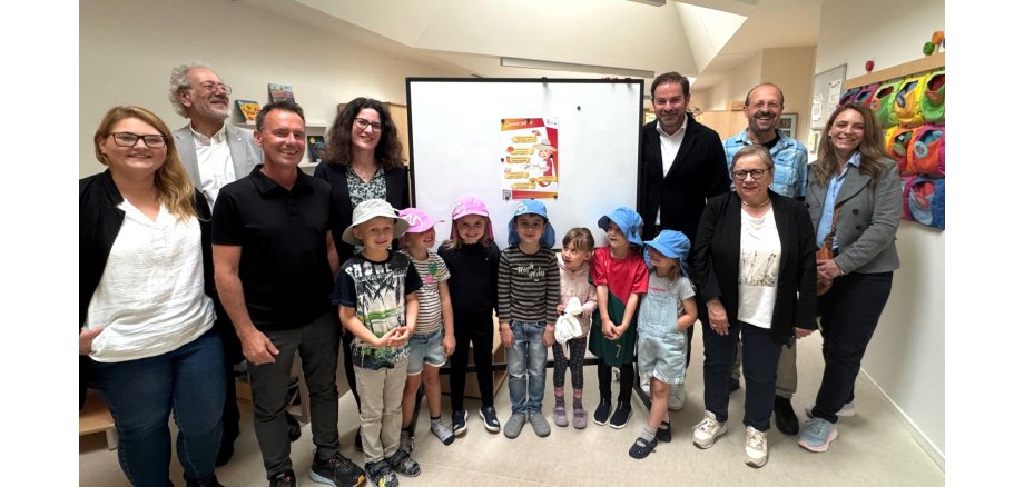 In einem Flur eines Kindergartens stehen 4 Frauen und 4 Männer und 7 Kinder, vor einem Board, an dem ein Plakat hängt. Die Kinder haben beige, rosa und blaue Sonnenhüte auf.