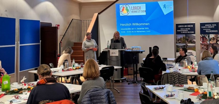 Design ohne Titel - 4 An vier rechtekigen Tischen, die mit Servietten, Mandarinen, Nüssen und Tannenzweigen dekoriert sind, sitzen mehrere Frauen. Vorn ist ein Rednerpult und ein Beamer auf einem Bistrotisch. Vorn stehen zwei Frauen am und neben dem Rednerpult. Hinter ihnen an der Wand ist das Titelbild einer Präsentation. Es sind zwei Rollups und Pinnwände aufgebaut. Im Hintergrund geht eine Treppe hoch in den nächsten Stock.