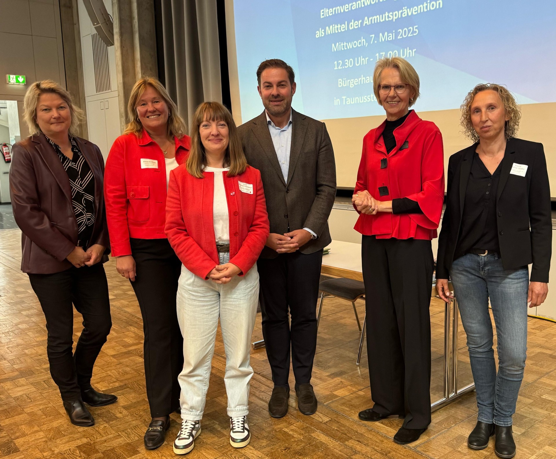 Landrat Zehner, steht mit fünf Frauen vor einem Tisch und einer Leinwand. Drei der Frauen tragen rote Blazer und weiße Namensschilder. 