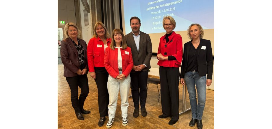 Landrat Zehner, steht mit fünf Frauen vor einem Tisch und einer Leinwand. Drei der Frauen tragen rote Blazer und weiße Namensschilder. 
