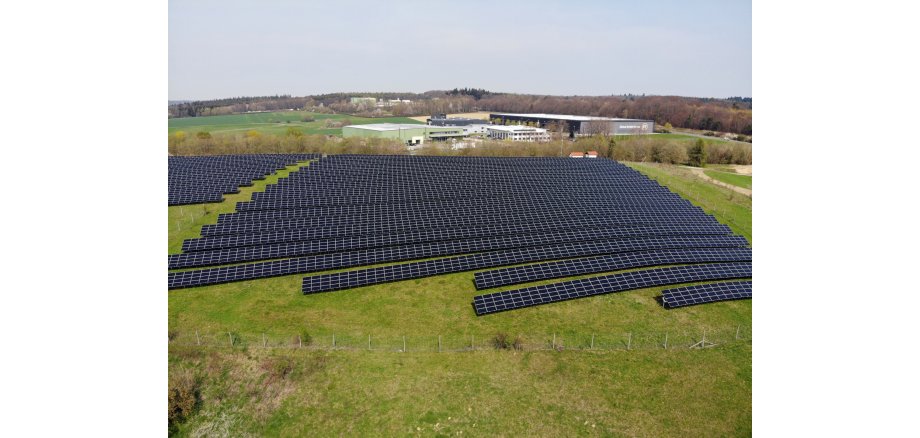 Aus der Luftperspektive wird ein Solarpark, der aus mindestens 19 Reihen und einer zweiten Fläche mit mindestens 11 Reihen Panelen besteht, dargestellt. Rund herum ist Wiese und im Hintergrund mehrere große Firmenhallen. Im Horizont ist Wald zu sehen. Der Himmel ist grau.