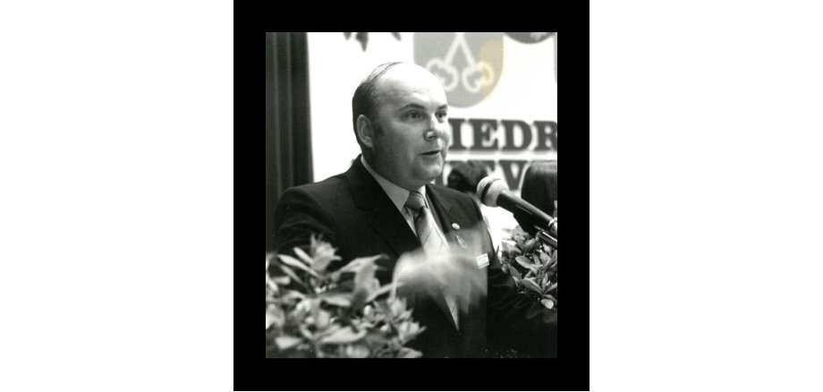 Ein Mann mit kurzen Haaren im dunkelm Anzug und Kravatte und weißem Hemd steht hinter einem Mikrofon. Links und rechts unten sind Blätter und im HIntergrund ein Vorhang und eine große Leinwand mit einem Wappen und Buchstaben. Das Bild ist in schwarz-weiß und hat einen schwarznen, breiten Rahmen.