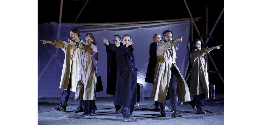 Auf einem grauen Boden stehen drei Männer und vier Frauen. Alle tragen helle oder dunkle, lange, geschlossene Mäntel und haben eine Aktenkoffer in der einen Hand. Die andere Hand ist ausgestreckt und mit dem Zeigefinger wird auf etwas gezeigt. Im Hintergrund ein dunkelblauer, durchsichtiger Banner, der an Stangen befestigt ist.