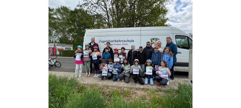 Vor einem weißen Kastenwagen mit der Beschriftung Jugendverkehrsschule RTK und dem Logo stehen 10 Kinder, zwei Männer und zwei Frauen und es knieen 7 Kinder davor auf dem Bürgersteig. Einige Kinder tragen Fahrradhelme und haben Flyer und Wimpel in der Hand.