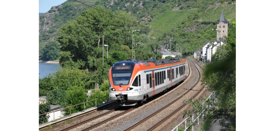 Ein Personenzug in orange und weiß fährt auf im Tal liegenden Schienen. Es ist ein Fluss zu erkennen und viele Bäume und ein aus Steinen gemauerter Kirchturm.