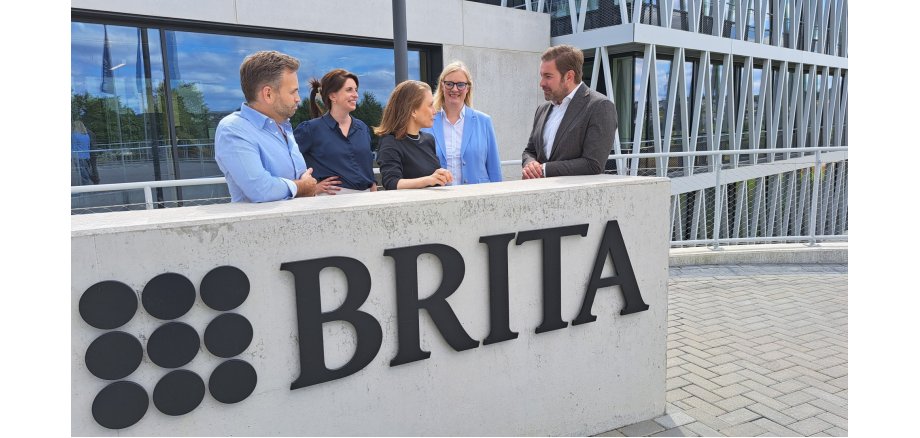 Hinter einer Betonwand, auf der das Firmenlogo von Brita geschrieben ist, stehen zwei Männer und drei Frauen. Dahinter ist eine breite Glasfront von Betonsteinen eingefasst und rechts ist ein großes Gebäude mit Stahlträgern als Dreiecken vor den Fenstern.