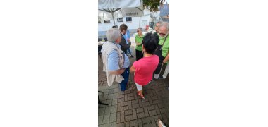 Auf einem gepflasterten Platz stehen unter einem Baum und einem Sonnenschirm zehn Personen und unterhalten sich.