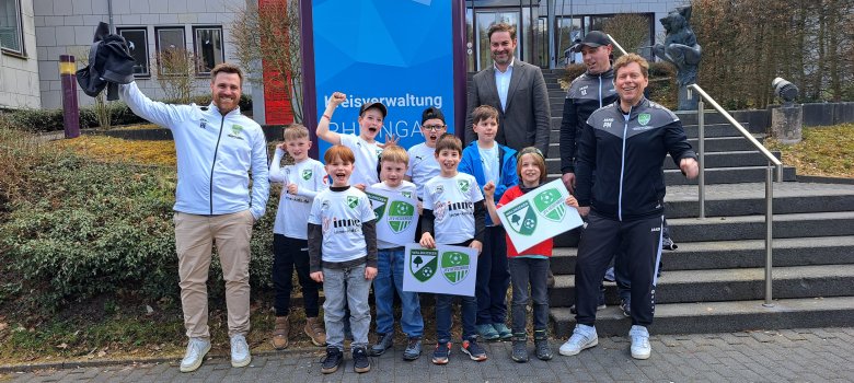 Vor einer langen Treppe und einem Gebäude mit vielen Fenstern der Kreisverwaltung und einem großem Schild mit Dreiecken mit unterschiedlichen Farben, stehen 8 Jungs mit Trikots und Schildern mit dem Logo ihres Fußballvereins. Ebenso steht Landrat Zehner im Anzug und drei Trainer in Trainingsjacken des Vereins dabei.
