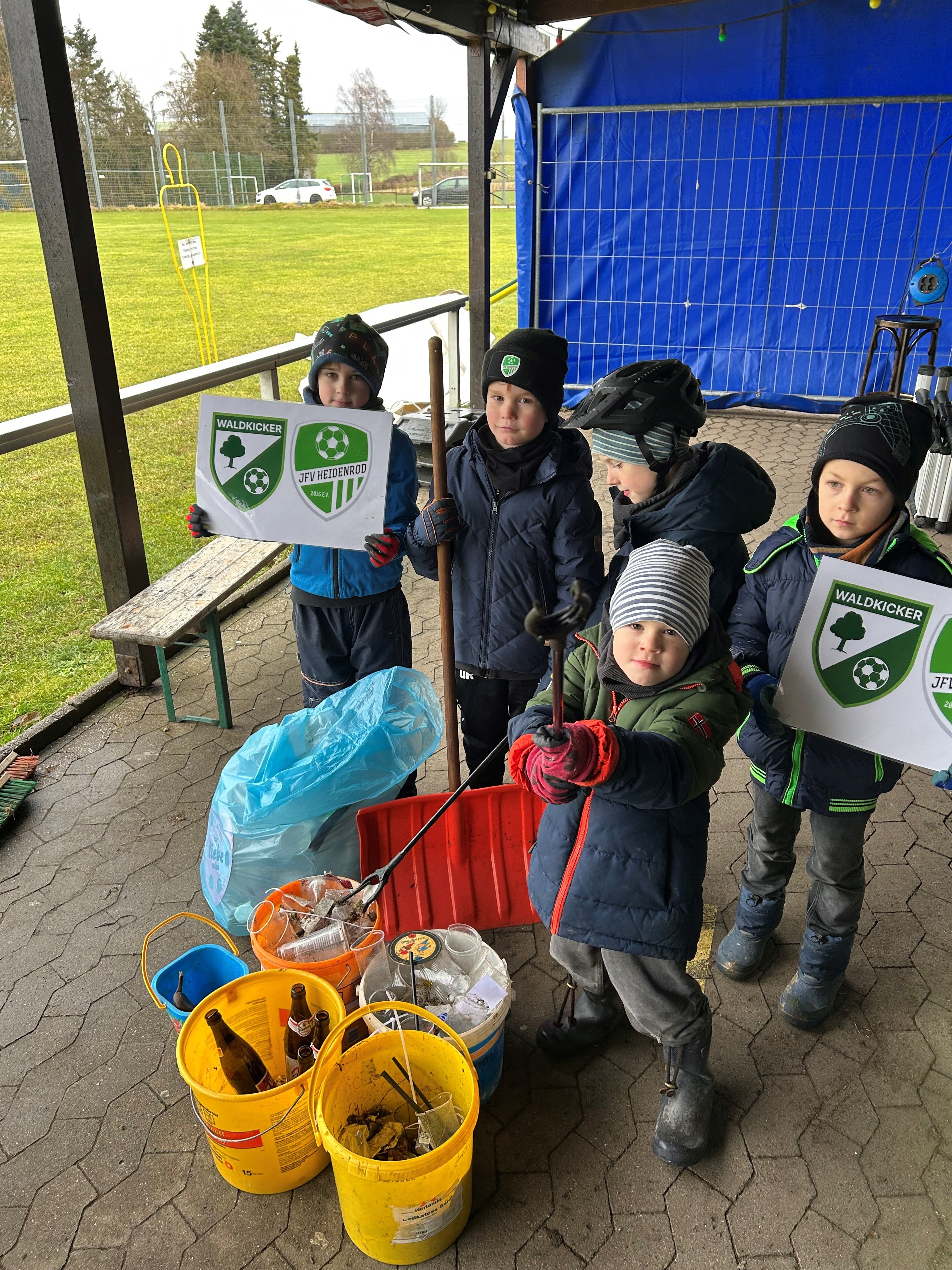Neben einer Wiese unter einem Unterstand stehen 5 Jungs mit Mützen und Anoraks und halten zwei Schilder mit dem Logo des Fussball-Vereins hoch. Vor ihnen stehen mehrere Eimer mit gesammeltem Müll.