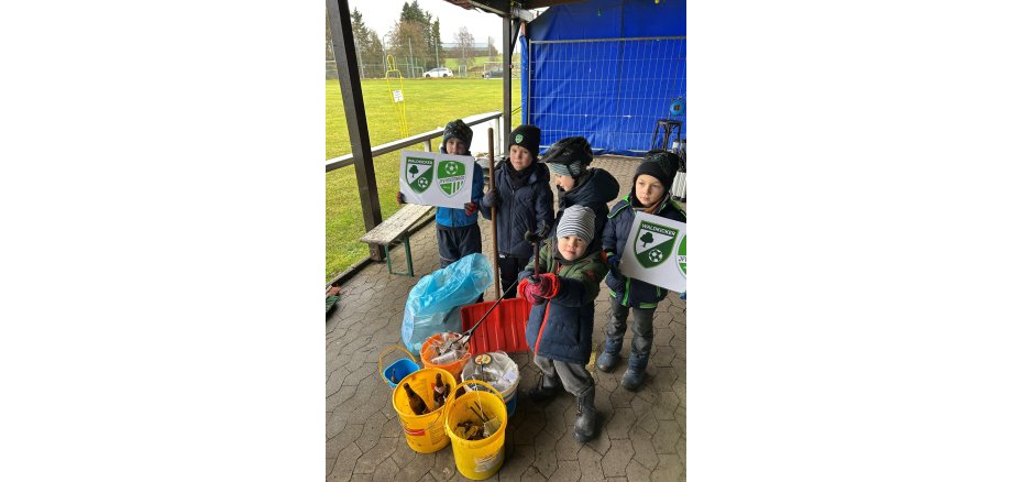 Neben einer Wiese unter einem Unterstand stehen 5 Jungs mit Mützen und Anoraks und halten zwei Schilder mit dem Logo des Fussball-Vereins hoch. Vor ihnen stehen mehrere Eimer mit gesammeltem Müll.