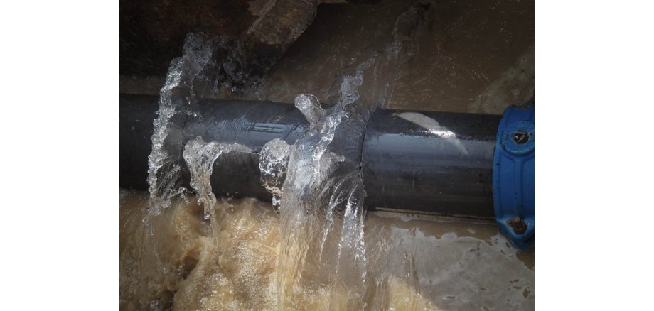 Aus einem großem grauem Rohr mit einer blauen Schelle tritt Wasser aus. Der Boden ist voller Wasser und bräunlich.