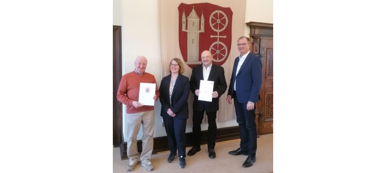 Vor der Fahne mit dem Kiedricher Wappen, dem Scharfenstein u nd dem Doppelrad in beige, auf rotem Untergrund, stehen v.l.n.r. Wilfried Ringel im orangenem Pullover und beiger Hose und Kerstin Merno, Georg Sohlbach, Bürgermeister Winfried Steinmacher, alle im dunklem Anzug mit weißem Hemd/Bluse. Herr Ringel und Herr Sohlbach haben Urkunden in der Hand.