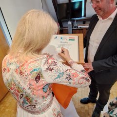 In einem Raum mit Holzparket und dunklen Holzbalken an den weißen Wänden und der weißen Decke steht ein Mann mit Glatze und heller Brille und weißem Hemd sowie dunklem Anzug hinter einem Gästebuch, in das sich eine Frau mit blonden, schulterlagen Haaren und weißem langärmeligem Kleid mit lachsfarbenene, braunen, hellblauen Ornamenten, einträgt. Die Frau steht mit dem Rücken zum Fotografen.