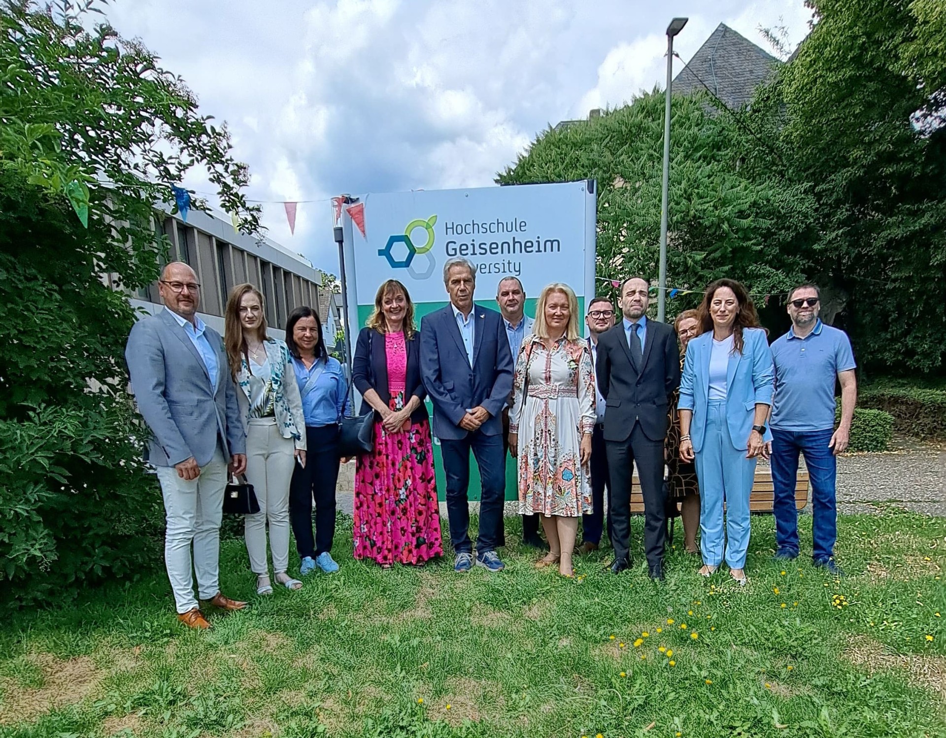 Sechs Männer und sechs Frauen stehen vor einem Schild mit der Aufschrift "Hochschule Geisenheim" auf einer Wiese. Im Hintergrund sind grüne Laubbäume und ein Gebäude mit Flachdach.