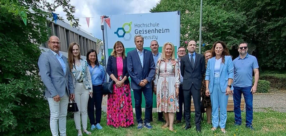 Sechs Männer und sechs Frauen stehen vor einem Schild mit der Aufschrift "Hochschule Geisenheim" auf einer Wiese. Im Hintergrund sind grüne Laubbäume und ein Gebäude mit Flachdach.