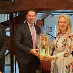 Links steht ein Mann mit heller Brille, Glatze, weißem Hemd und dunkelgrauem Anzug. In der MItte steht ein Mann im weißen Hemd mit rosa farbener Krawatte und dunklem Anzug mit einem Wein-Geschenk in einer Holzkiste, welches in Klarsichtfolie gepackt ist. Rechts davon steht eine Frau mit blonden, schulterlagen Haaren, die ein weißes, langärmeliges Kleid mit lachsfarbenen, braunen, hellblauen Ornamenten trägt und das Geschenk in den Händen hält.