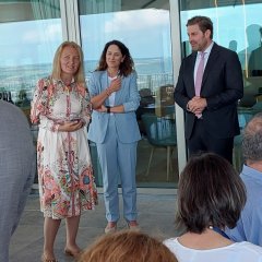 Vor großen Glas-Schiebetüren steht rechts ein Mann im weißen Hemd mit rosa farbener Krawatte und dunklem Anzug. In der Mitte steht eine Frau mit braunen, gelockten, langen Haaren in hellblauem Hosenanzug und weißem Shirt. Links davon eine Frau mit blonden, schulterlagen Haaren, die ein weißes, langärmeliges Kleid mit lachsfarbenen, braunen, hellblauen Ornamenten trägt. 