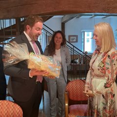 Hinter einem Tisch mit einer gelben viereckigen Tischdecke und Namensschild, einem Wasserglas, Zetteln, Mappen und Stiften, steht ein Mann im weißen Hemd mit rosa farbener Krawatte und dunklem Anzug mit einem Geschenk in den Händen welches in Klarsichtfolie gepackt ist. Rechts davon eine Frau mit blonden Schulterlagen Haaren, die ein weißes, langärmeliges Kleid mit lachsfarbenen, braunen, hellblauen Ornamenten trägt. Dazwischen steht im Hintergrund eine Frau mit braunen langen Haaren in hellblauem Hosenanzug und weißem Shirt.