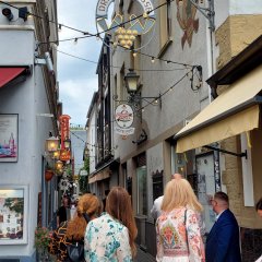 Blick auf eine schmale Gasse, die mit dem Schild "Drosselgasse" und Weintrauben dekoriert ist. Auf dem Bild stehen drei Frauen und zwei Männer mit dem Rücken zum Fotografen, am Eingang der Gasse auf Kopfsteinpflaster.