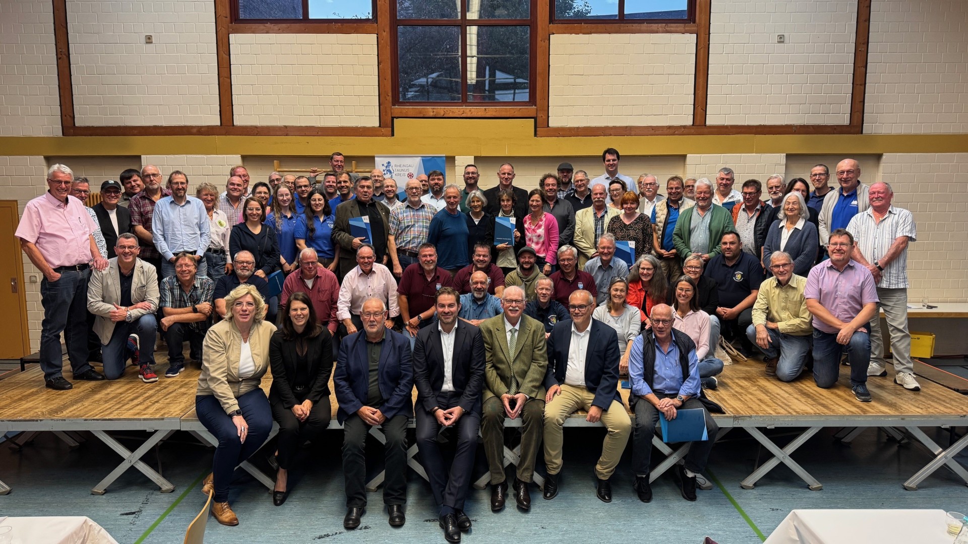 Auf einer großen Bühne, die auf gekreuzten Stelzen steht, stehen, sitzen und knien ca. 80 Personen. Die Bühne steht vor einer gemauerten beigen Wand mit braunen Streifen und Fenstern.