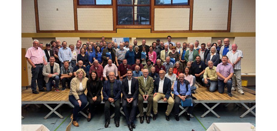 Auf einer großen Bühne, die auf gekreuzten Stelzen steht, stehen, sitzen und knien ca. 80 Personen. Die Bühne steht vor einer gemauerten beigen Wand mit braunen Streifen und Fenstern.