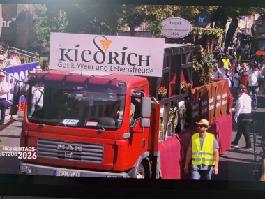 Ein roter LKW, der auf der Fahrerkabine ein großes weißes Schild mit der Aufschrift "Kiedrich" hat. Er ist rot geschmückt und an den Seiten sind ovale Holzscheiben mit den Namen der Weinlagen. Auf dem LKW sind rote Blumen und eine Pergola aus dunklen Holzbalken. Neben dem LKW läuft ein Mann mit Bart, Sonnenbrille, Sonnenhut und neongelber Warnweste. Hinter dem Wagen ist eine Fußgruppe.